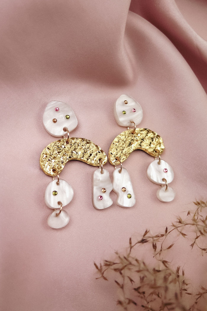 A pair of boho chic dangle earrings featuring gold textured arches and white, marble-like pieces, adorned with small colored gems, rhinestones, and displayed on soft pink fabric with dried flowers beside them.