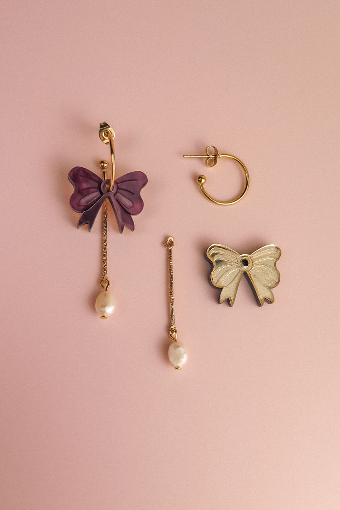 Three mismatched earrings on a pink background: a gold-plated hoop earring, a purple bow with a dangling freshwater pearl, and a white bow edged in gold attached to a bar and pearl—perfect for creating your own interchangeable dangle earring look.