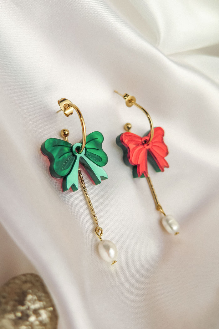 Two gold-plated c-hoop earrings with bow-shaped charms, one green and one red, each featuring a freshwater pearl dangling from a gold chain—stylish interchangeable dangle earrings displayed on white fabric.