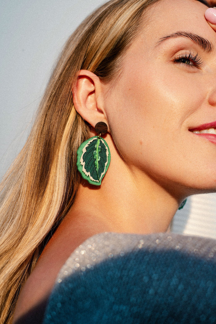 A woman with long blonde hair wears a large green leaf-shaped earring, reminiscent of nature-inspired earrings like the Calathea Medallion Earrings, and smiles softly in bright sunlight.