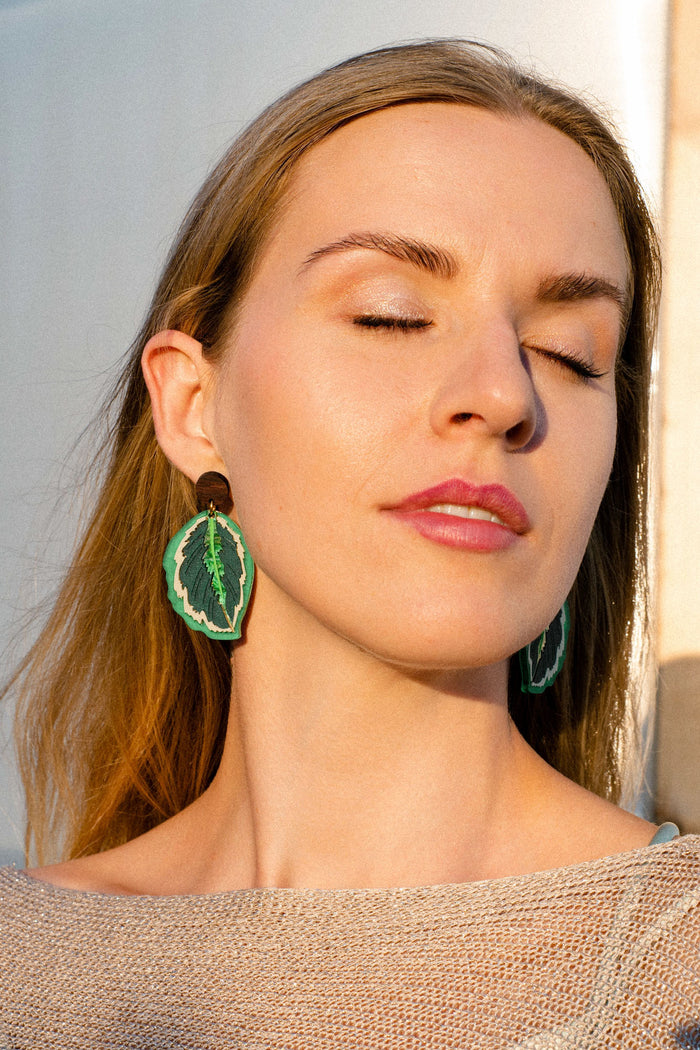 A woman with long hair and closed eyes wears large green, nature-inspired Calathea Medallion Earrings—handcrafted statement jewelry—standing in sunlight and wearing a sheer, light-colored top.