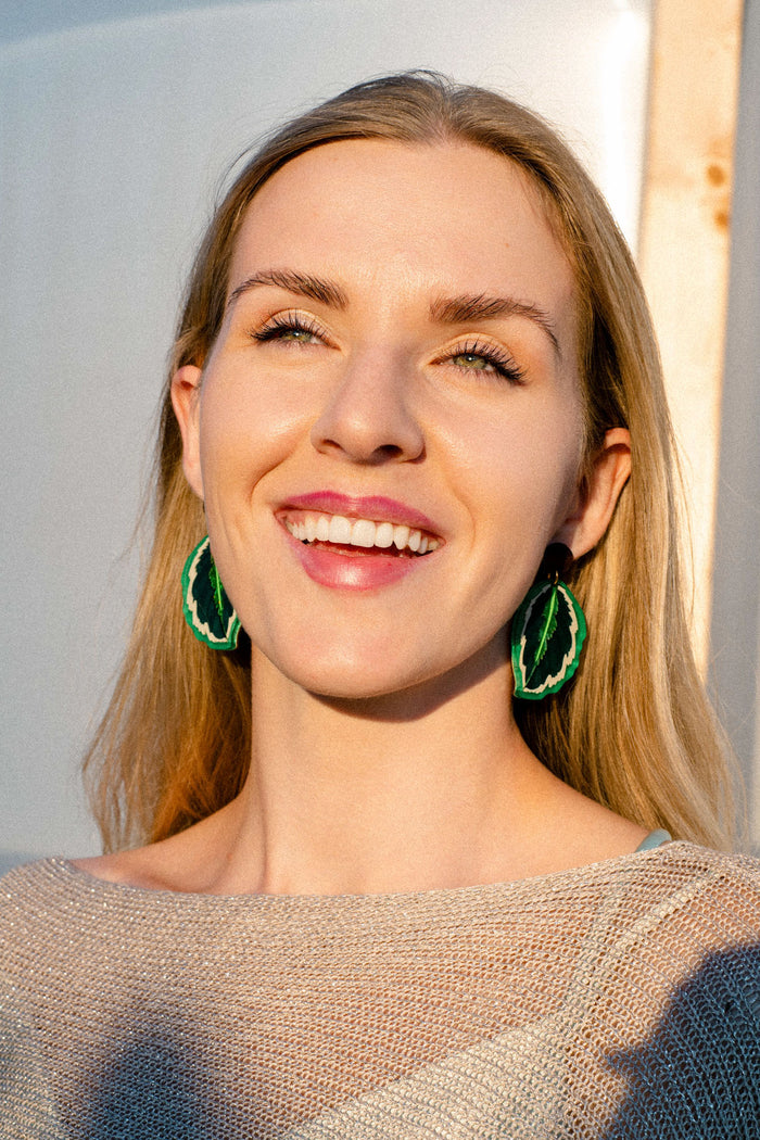 A smiling woman with long blonde hair and green leaf-shaped Calathea Medallion Earrings stands in sunlight, wearing a beige top. Her handcrafted statement jewelry adds a bold, nature-inspired touch to her look.