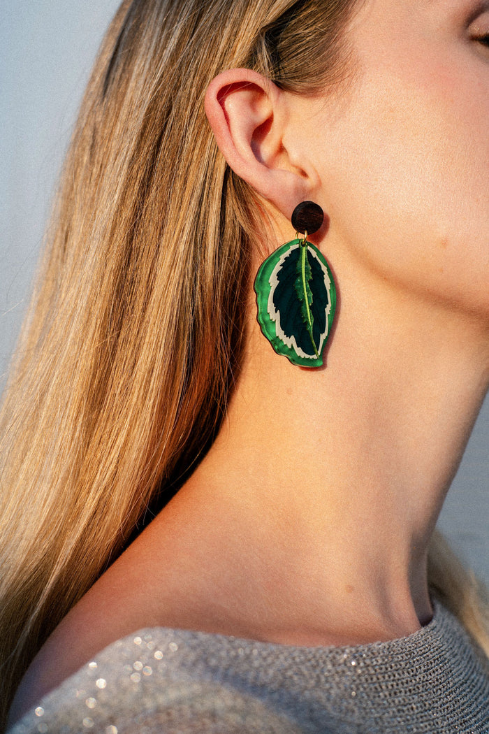 A close-up of a person with long blonde hair wearing a large, leaf-shaped Calathea Medallion Earring featuring green and black colors, and a sparkly light-colored top. These nature-inspired earrings beautifully showcase handcrafted statement jewelry at its finest.