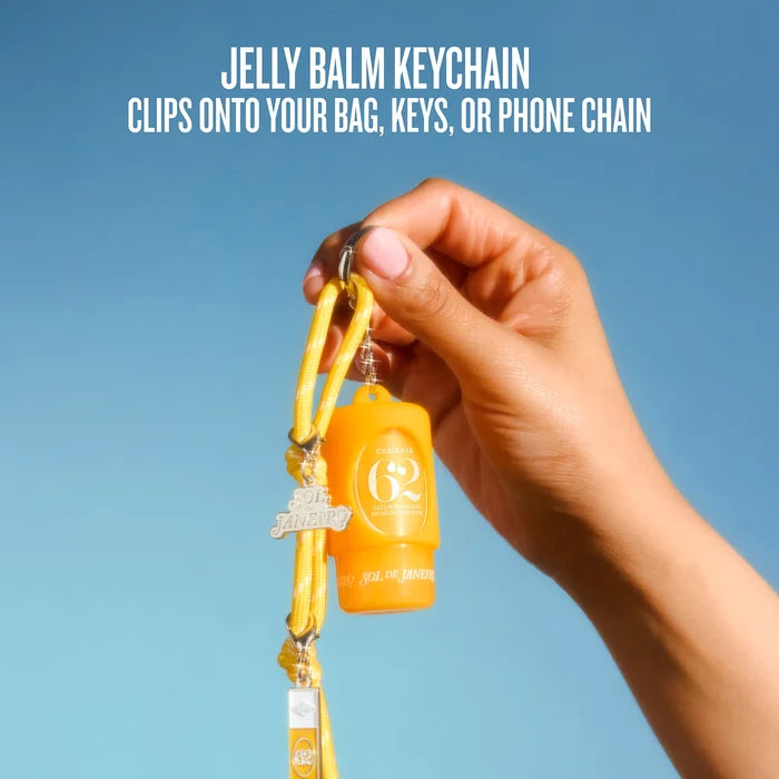 A hand holding a yellow and orange 'G2' jelly balm keychain with a yellow cord and silver carabiner, set against a clear blue sky. White text at the top reads 'JELLY BALM KEYCHAIN CLIPS ONTO YOUR BAG, KEYS, OR PHONE CHAIN'.
