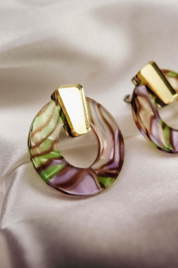 Close-up of a round, marbled earring made from acrylic glass with pink, green, and purple swirls, featuring a shiny gold geometric accent on top. This hypoallergenic piece rests elegantly on soft, cream-colored satin fabric.