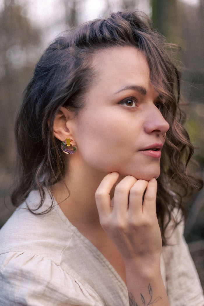 A woman with wavy brown hair and a light beige blouse looks to the side outdoors, resting her chin on her hand. She wears colorful hypoallergenic Minimalist Circle Stud Earrings and has a subtle tattoo on her wrist.