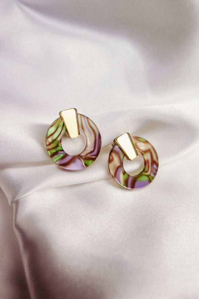 A pair of minimalist circle stud earrings featuring gold geometric tops and marbled, multicolored acrylic glass bases in pink, green, and beige tones, displayed on white satin fabric. Hypoallergenic for comfortable wear.