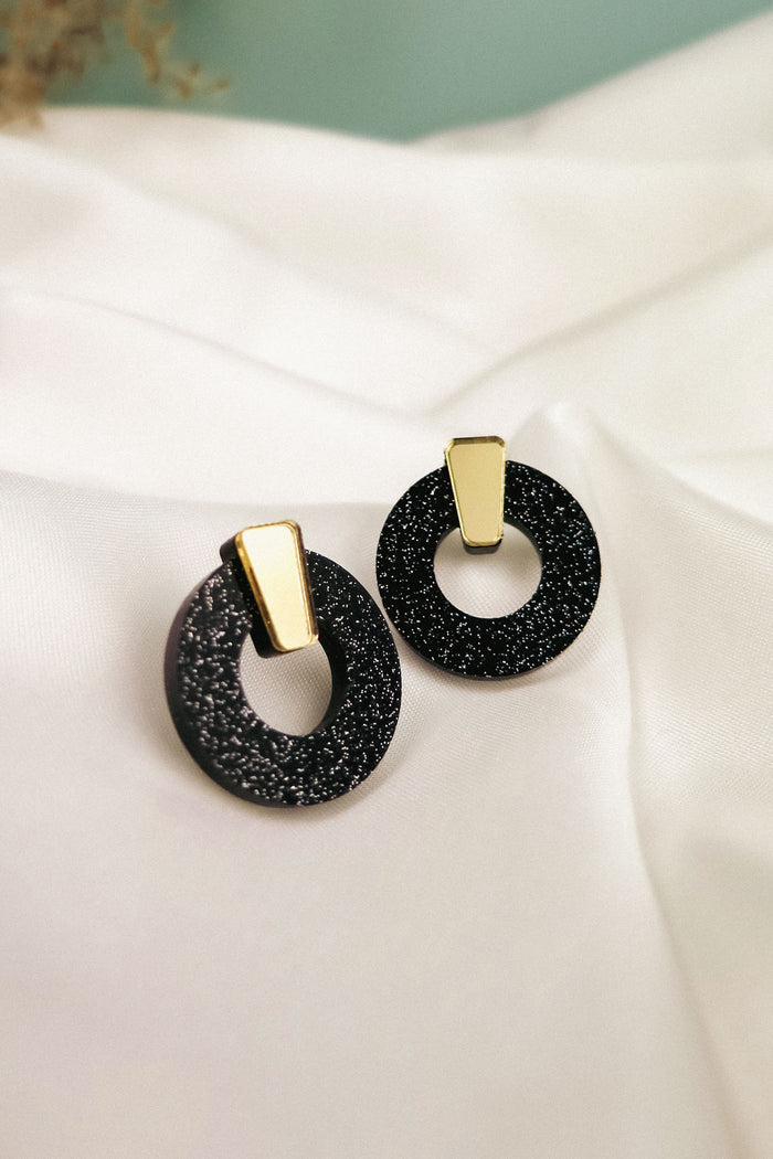 A pair of stylish Minimalist Circle Stud Earrings featuring black glittery acrylic glass hoops with gold rectangular accents, displayed on a white satin fabric background. Hypoallergenic for comfortable, all-day wear.