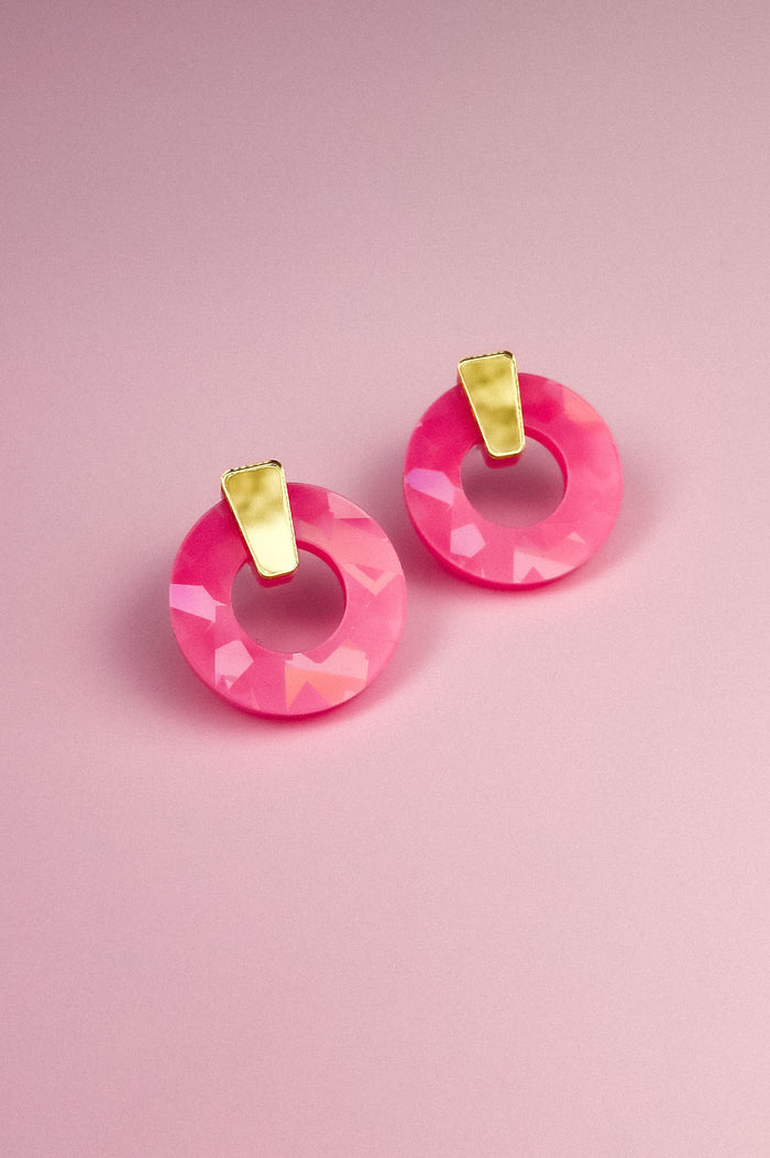 A pair of Minimalist Circle Stud Earrings featuring circular, faceted pink acrylic glass with elegant gold accents, displayed on a soft pink background.