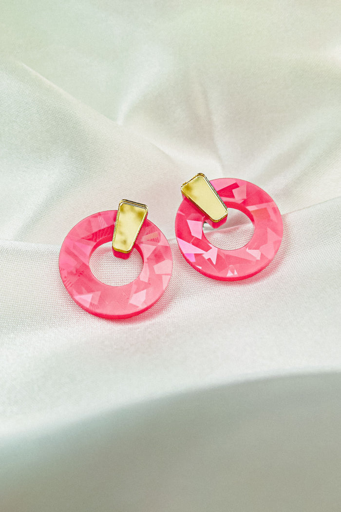 A pair of Minimalist Circle Stud Earrings featuring large, faceted pink acrylic glass hoops and small, irregular yellow rectangular studs, displayed on a soft white satin fabric background.