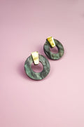A pair of hypoallergenic Minimalist Circle Stud Earrings, featuring round green marbled acrylic glass and gold rectangular studs, displayed on a pink background.