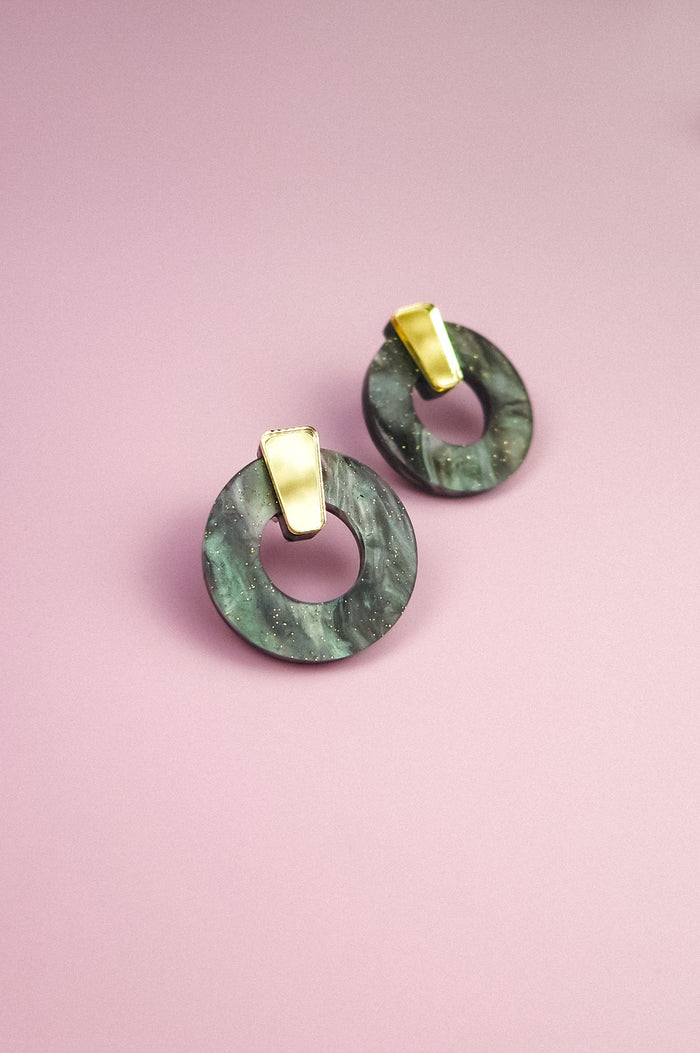 A pair of hypoallergenic Minimalist Circle Stud Earrings, featuring round green marbled acrylic glass and gold rectangular studs, displayed on a pink background.