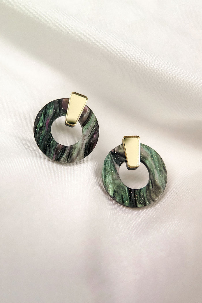 A pair of round, marbled green and black acrylic glass earrings with gold triangular studs, displayed on a soft white fabric background. These hypoallergenic Minimalist Circle Stud Earrings combine style and comfort beautifully.