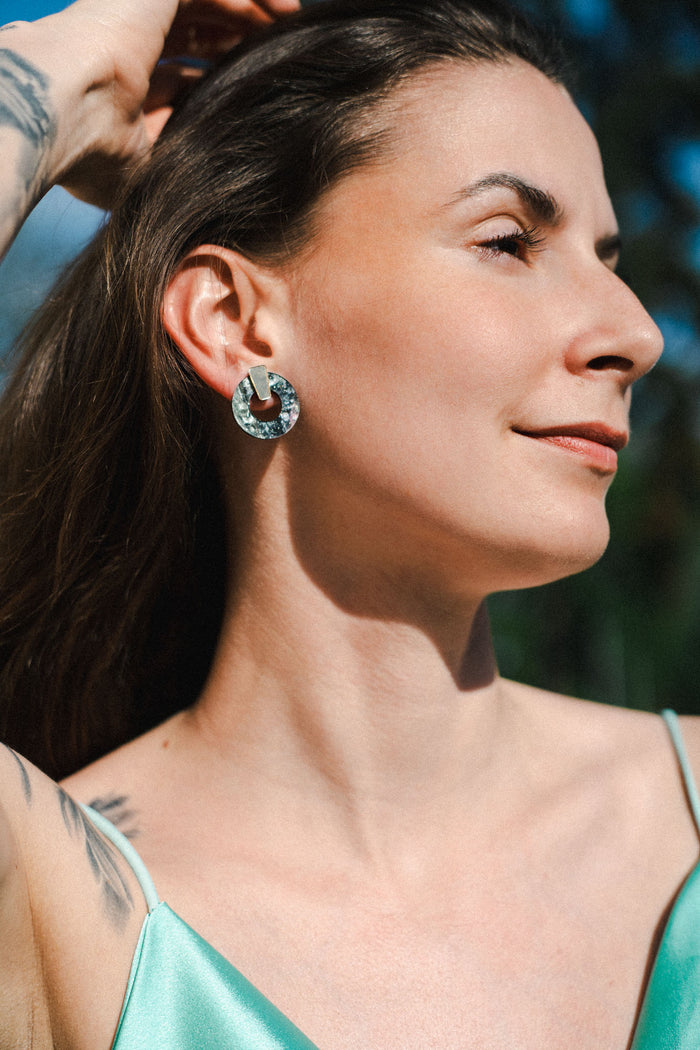 A woman with long brown hair and a tattoo on her arm poses outdoors in sunlight, wearing a green satin top and minimalist circle stud earrings, looking off to the side and smiling slightly.