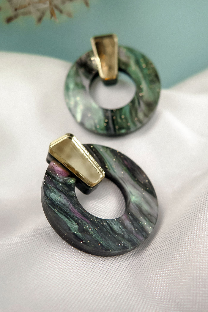 Minimalist Circle Stud Earrings crafted from acrylic glass feature marbled green, black, and purple hues with gold rectangular accents. They rest on white textured fabric, against a softly blurred light blue backdrop. Hypoallergenic for sensitive ears.