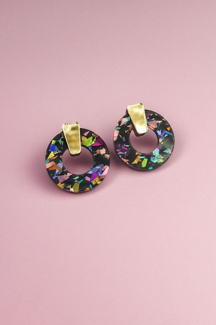 A pair of minimalist circle stud earrings with multicolored, iridescent patterns crafted from acrylic glass and gold rectangular clips, displayed on a pink background.