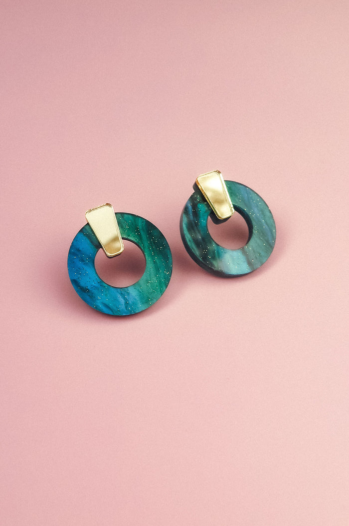 Minimalist Circle Stud Earrings crafted from blue-green marbled acrylic glass with gold triangular tops, displayed on a pink background. These hypoallergenic earrings offer both style and comfort.