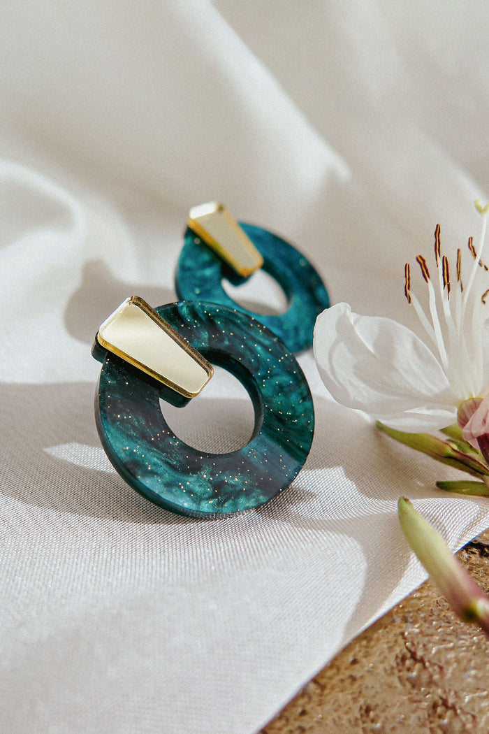 Two minimalist circle stud earrings with a marbled teal design and small gold accent, crafted from hypoallergenic acrylic glass, are displayed on a white cloth next to a white flower with pink details.