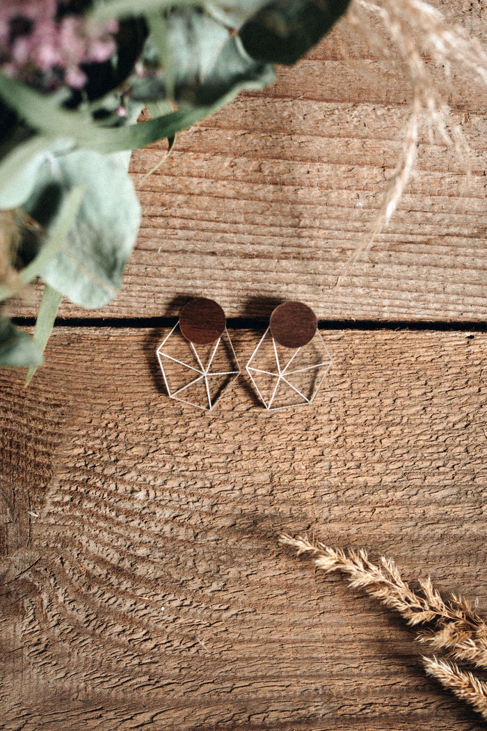 A pair of geometric earrings with wooden circular tops and silver wire shapes below, displayed on a rustic wooden surface with green leaves and dried wheat in the corners, exuding minimalist elegance.