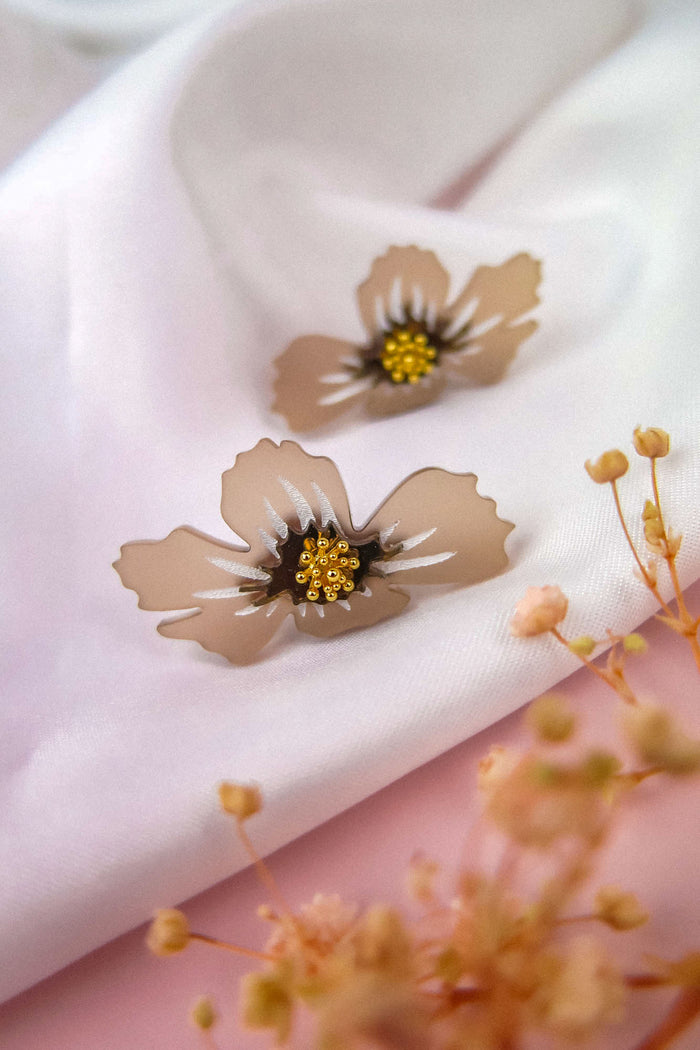 Two beige, translucent flower-shaped handmade earrings with yellow and black centers rest on a white fabric background, with blurred dried flowers in the foreground.