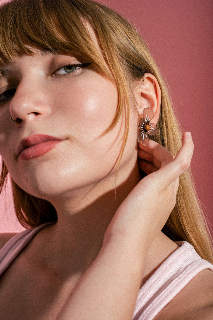 A woman with light brown hair and glowing skin poses against a pink background, showing off a delicate acrylic flower earring while wearing a light pink sleeveless top.