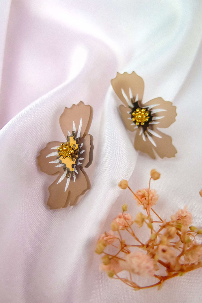 Two beige acrylic flower earrings with yellow beaded centers rest on soft white fabric, alongside small dried pink flowers. These handmade earrings capture a delicate, feminine charm perfect for any occasion.