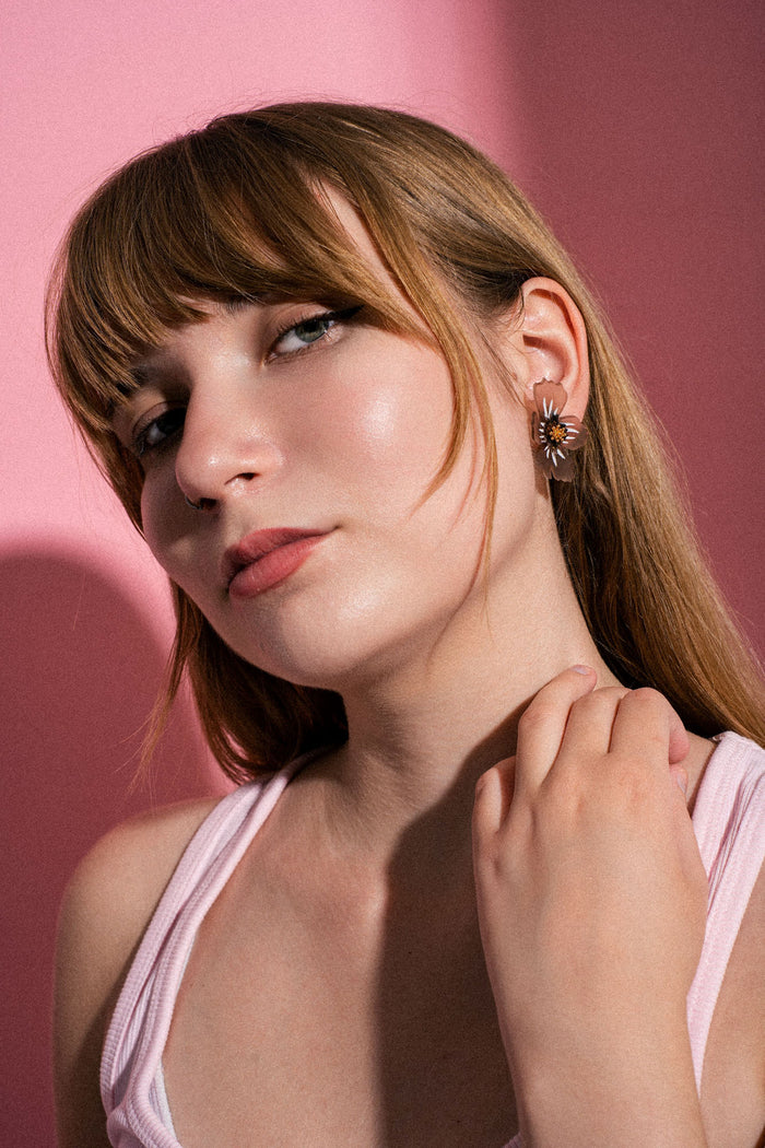 A woman with straight light brown hair and bangs poses against a pink background, wearing a pink top and handmade acrylic flower earrings, gazing confidently at the camera with her hand near her neck.