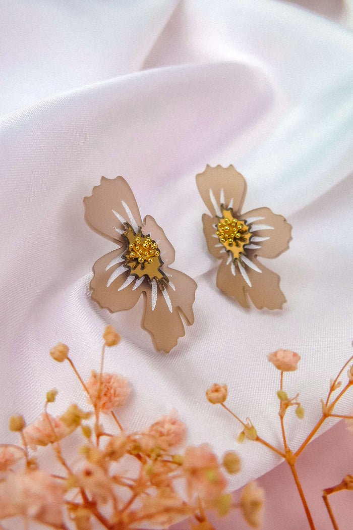 Beige acrylic flower earrings with yellow and white centers rest on soft, pale pink fabric. Delicate dried flowers are visible in the foreground, adding a gentle, elegant touch to the composition and highlighting the unique charm of these handmade earrings.
