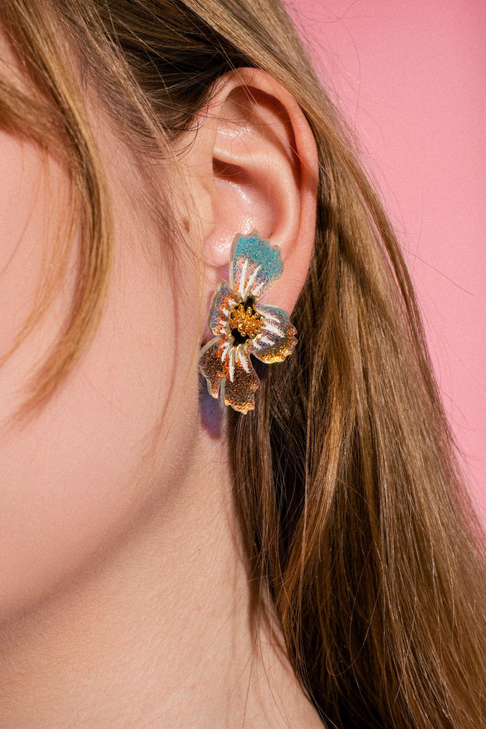 Close-up of a woman’s ear wearing a large, colorful acrylic flower earring with gold, blue, purple, and pink tones. The frosted earrings catch the light beautifully against the pink background, while her light brown hair partially covers her ear.