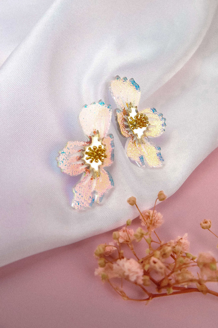 Two iridescent, handmade acrylic flower earrings with gold centers rest on white satin fabric, with small dried flowers placed nearby on a pink background.
