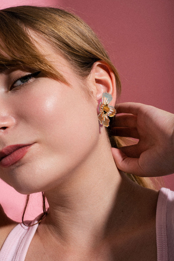 A close-up of a woman with light skin and brown hair, wearing a large, frosted acrylic flower earring and touching her neck, set against a pink background.