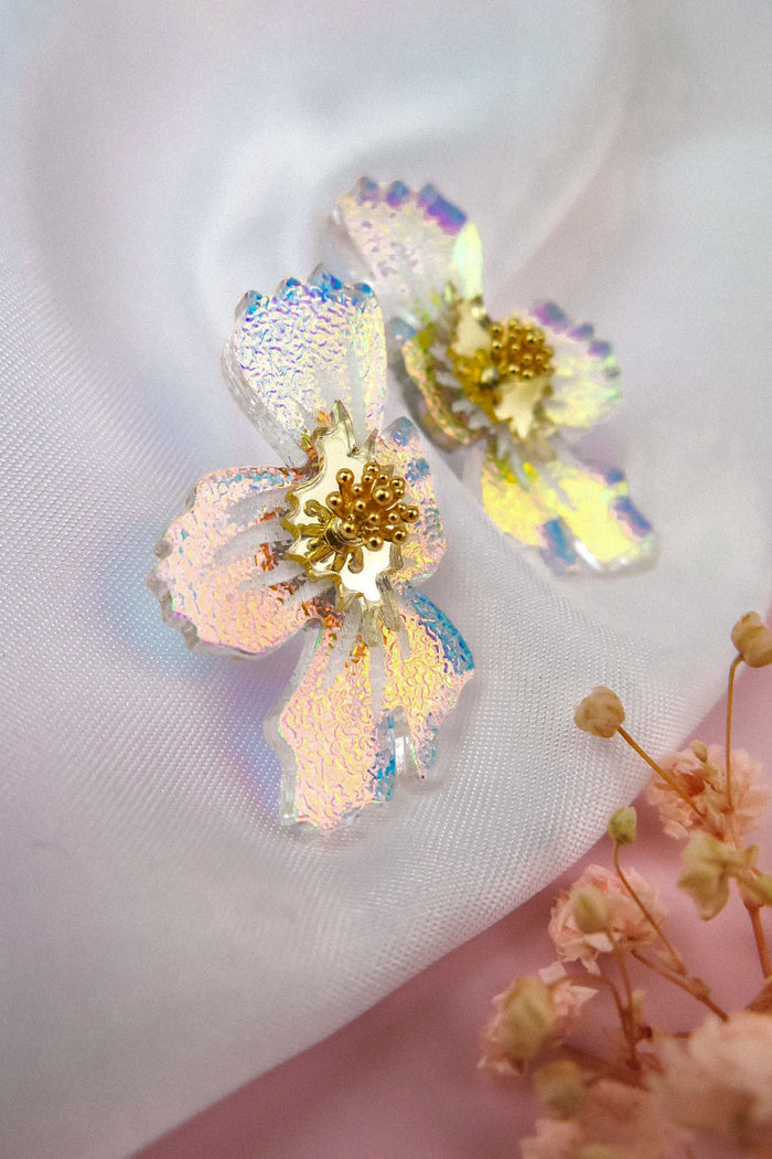 Iridescent, frosted acrylic flower earrings with textured petals and gold centers displayed on white fabric, with delicate dried flowers in the corner—handmade earrings that beautifully capture a touch of nature’s elegance.