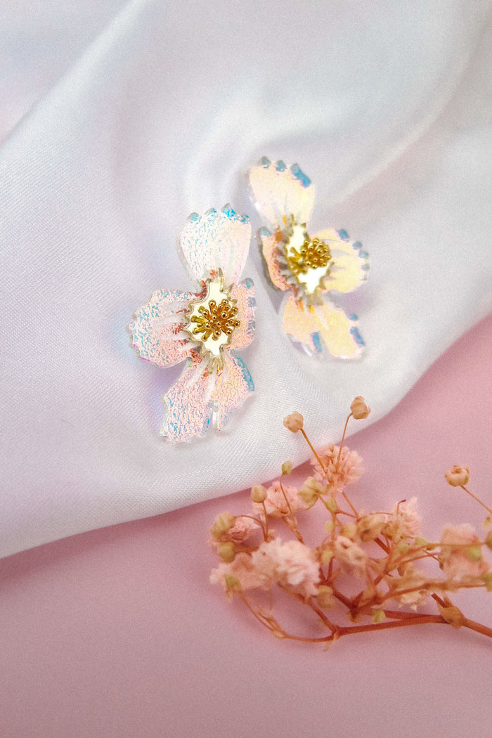 Iridescent acrylic flower earrings with yellow centers rest on white fabric, accompanied by small dried pink flowers on a pastel pink surface. These frosted earrings showcase a delicate, handmade elegance that brightens any look.