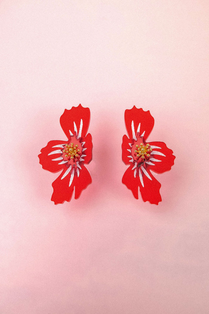 Two red, stylized acrylic flower earrings with white and yellow details are displayed on a light pink background.