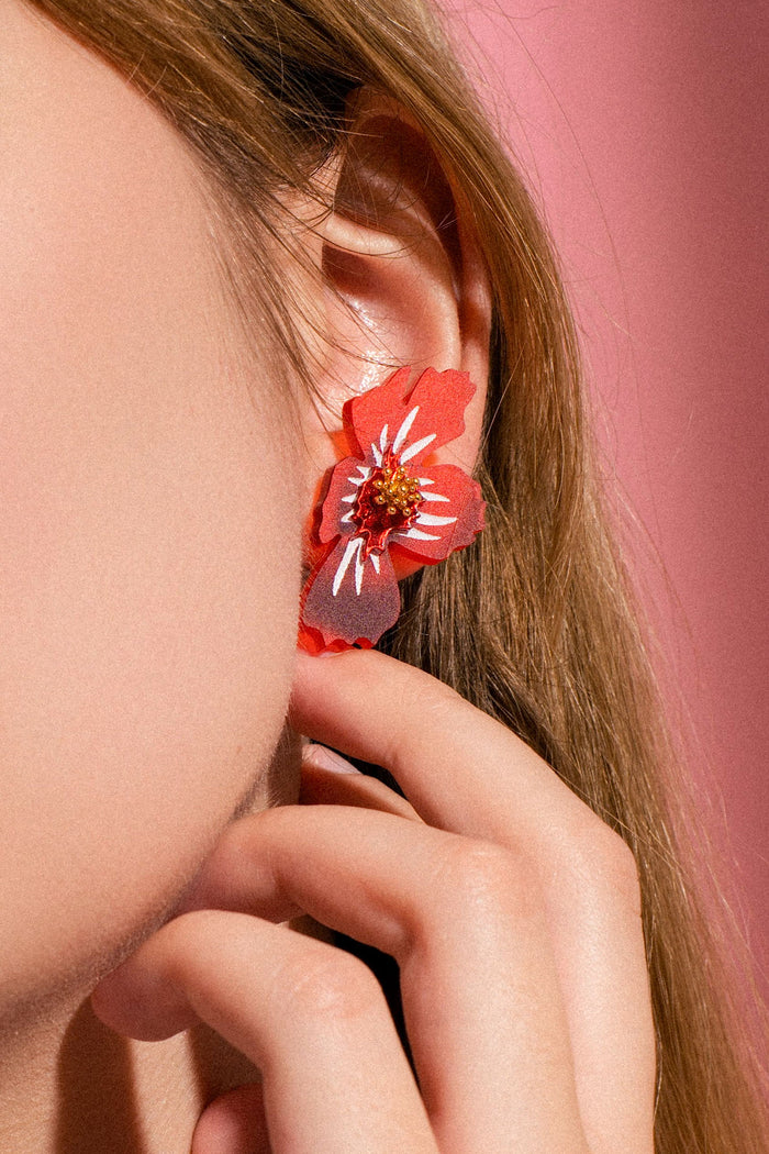A close-up of a person’s ear wearing a colorful, acrylic flower earring. The person touches their face with their fingers, and the background is pink, highlighting the playful charm of these handmade earrings.