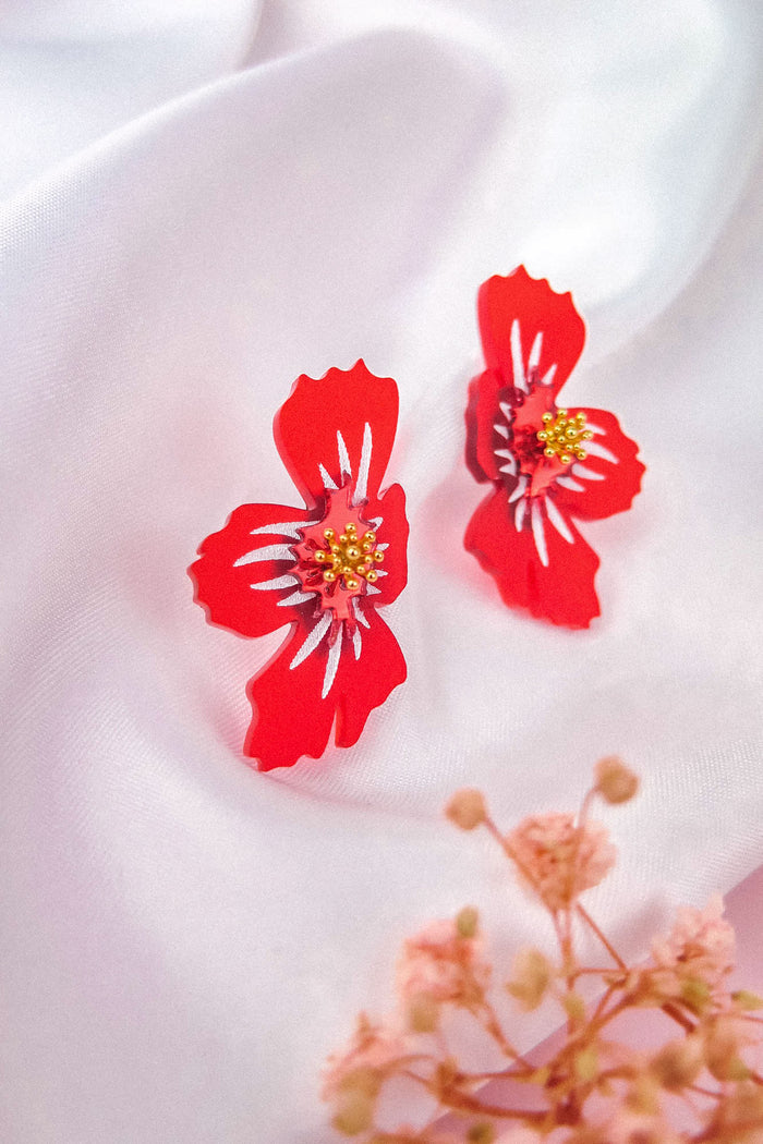 Red acrylic flower earrings with yellow and white accents rest on a soft white fabric, with a blurred sprig of small pink flowers in the foreground. These handmade earrings feature a delicate frosted finish that adds an elegant touch.