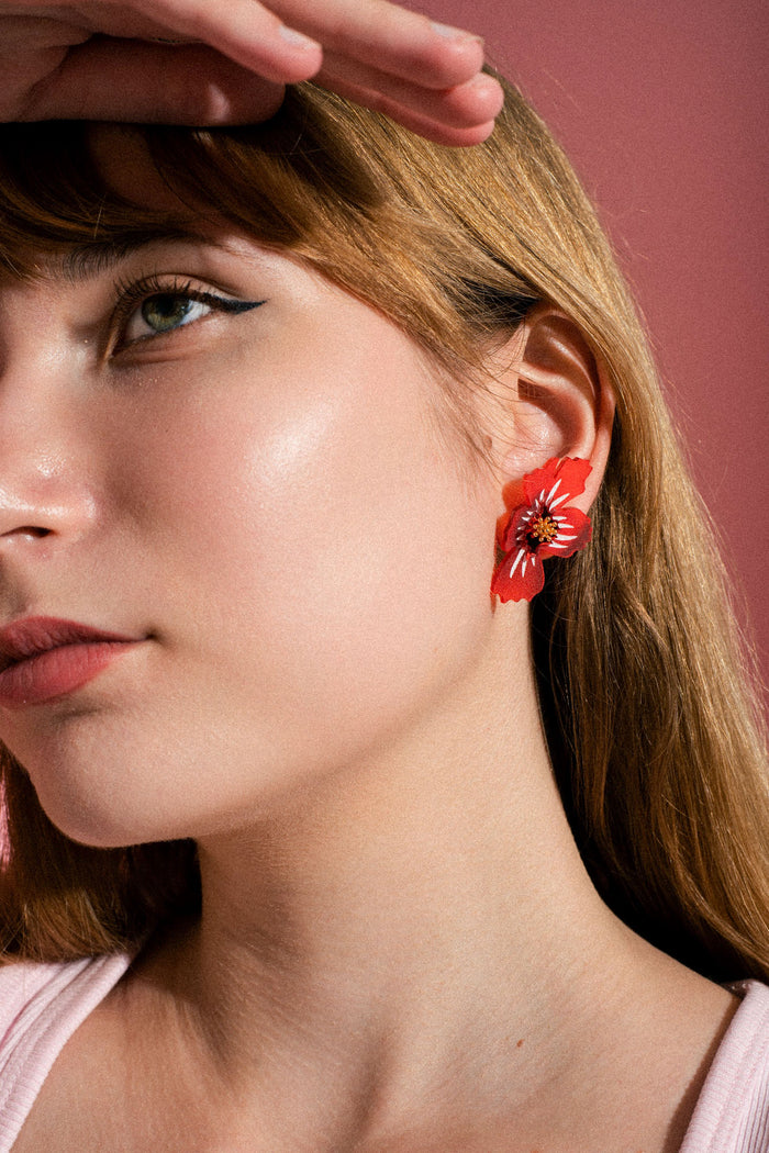 Close-up of a woman with light brown hair, wearing handmade acrylic flower earrings, looking up with her hand resting on her forehead, against a pink background.