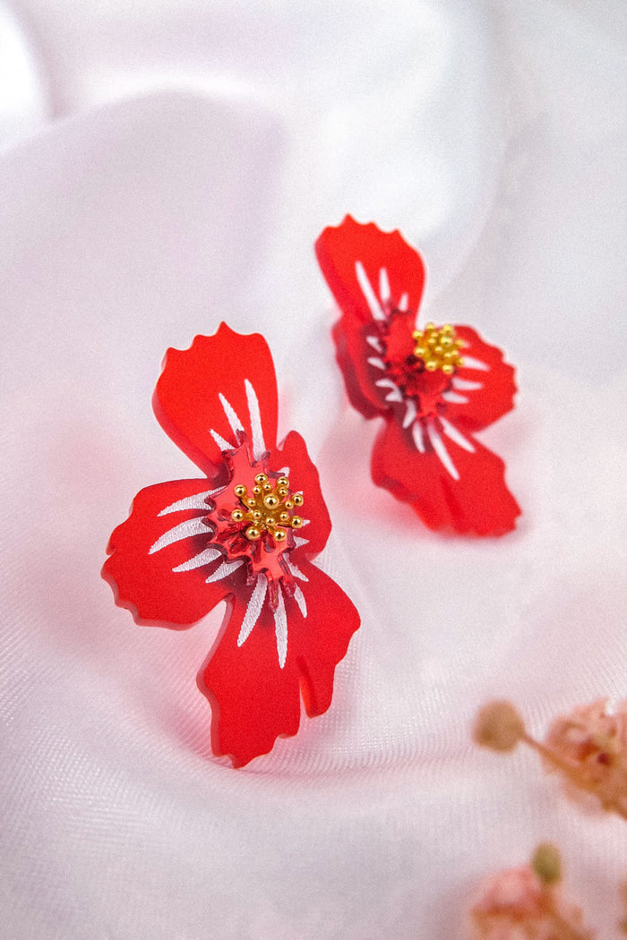 Two red acrylic flower earrings with yellow bead centers rest on a soft white fabric, their frosted petals catching the light, while a small cluster of pinkish dried flowers is blurred in the foreground.