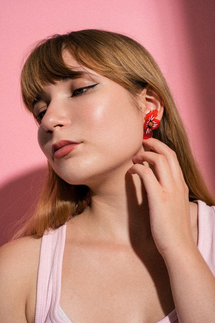 A woman with long light brown hair poses against a pink background, wearing a pink top and a pair of striking acrylic flower earrings, gently touching her neck with one hand.
