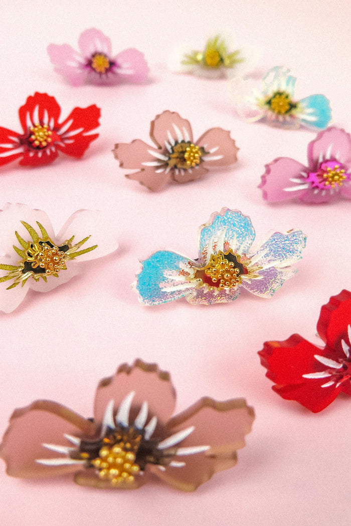 Assorted colorful acrylic flower embellishments with gold bead centers, reminiscent of delicate acrylic flower earrings, are arranged on a light pink background.