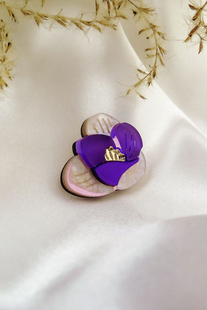 A purple and cream flower-shaped brooch, reminiscent of a Purple Crocus Flower Lapel Pin with gold detailing, rests on smooth white satin fabric, surrounded by delicate dried grass—perfect for nature lovers.