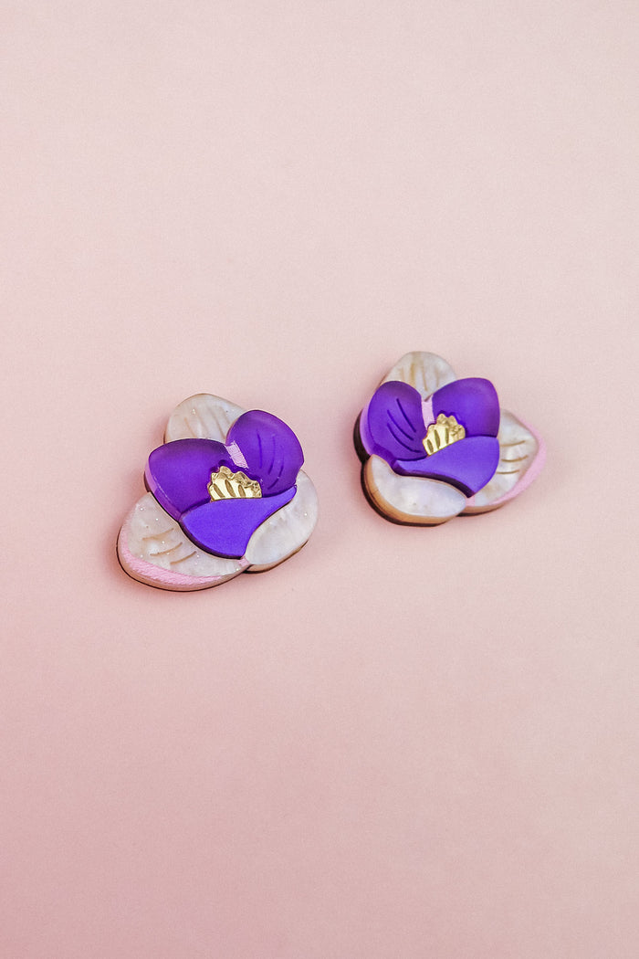 Crocus Stud Earrings shaped like purple flowers with yellow centers, set against a light pink background. The petals are layered, hand-painted flowers with a delicate, handmade appearance and are allergy-free.