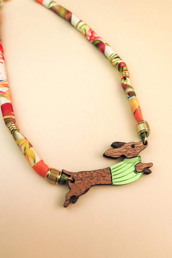 A piece of handcrafted jewelry featuring a colorful, patterned fabric cord and a wooden pendant shaped like a leaping rabbit in a green shirt. Made with recycled materials, set against a soft beige background.
