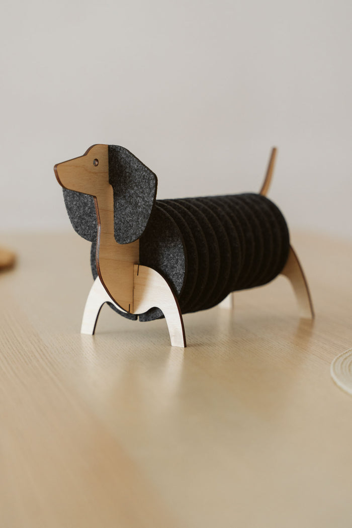 A coaster holder shaped like a dachshund dog, with a wooden head, tail, and legs, displays stacked round black felt coasters as its body, sitting on a light wood surface.