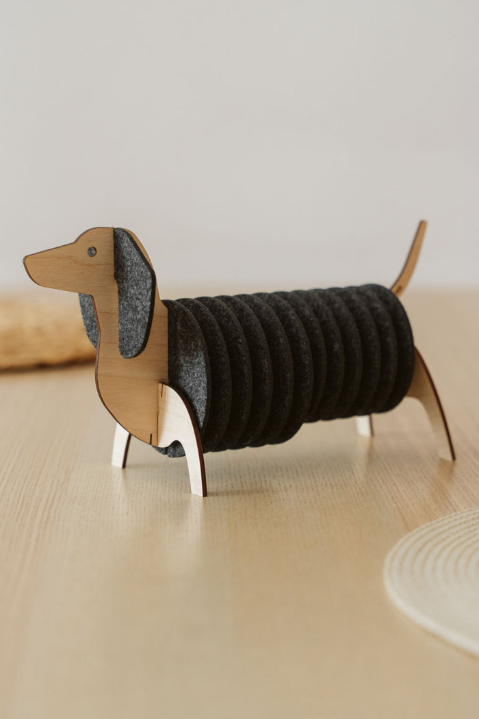 A wooden dachshund-shaped holder with dark felt coasters stacked as the dog’s body, placed on a light-colored table. Perfect for fans of Dachshund coasters and charming decor.