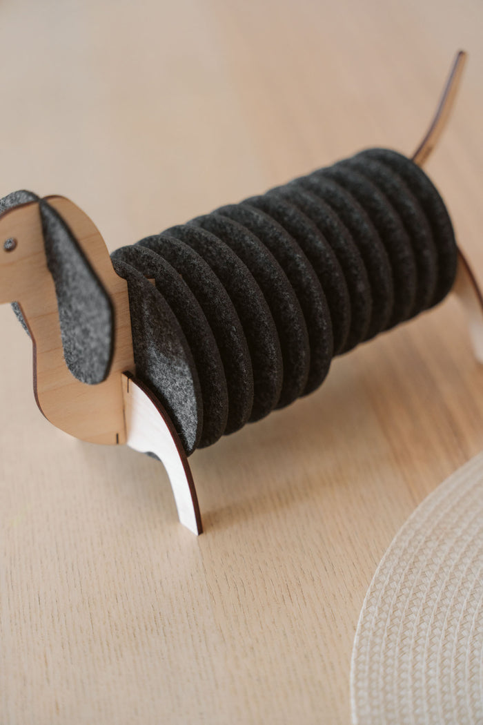 A napkin holder shaped like a dachshund dog, with a wooden head, legs, and tail, and black felt body segments holding napkins, sits on a light wooden table alongside matching dachshund coasters.