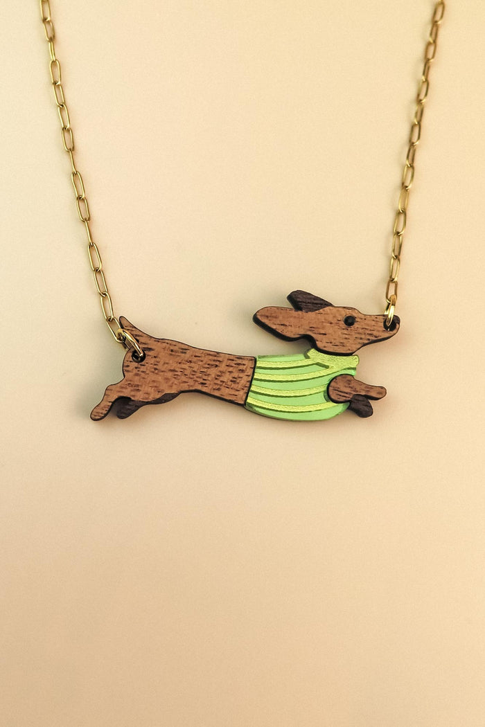 A necklace with a gold chain features a pendant of a brown Dachshund wearing a colorful sweater, depicted in a running pose against a beige background.