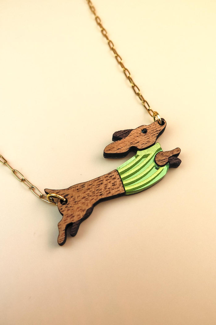 A gold chain necklace featuring a wooden Dachshund pendant in a colorful sweater, crafted from sustainable wood. The leaping pup wears green and yellow stripes against a light cream background.