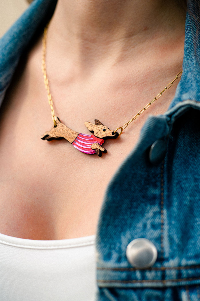 A close-up of a person wearing a gold necklace with a handcrafted Dachshund pendant in a pink striped sweater, made from sustainable wood. The person is also wearing a white top and a blue denim jacket.