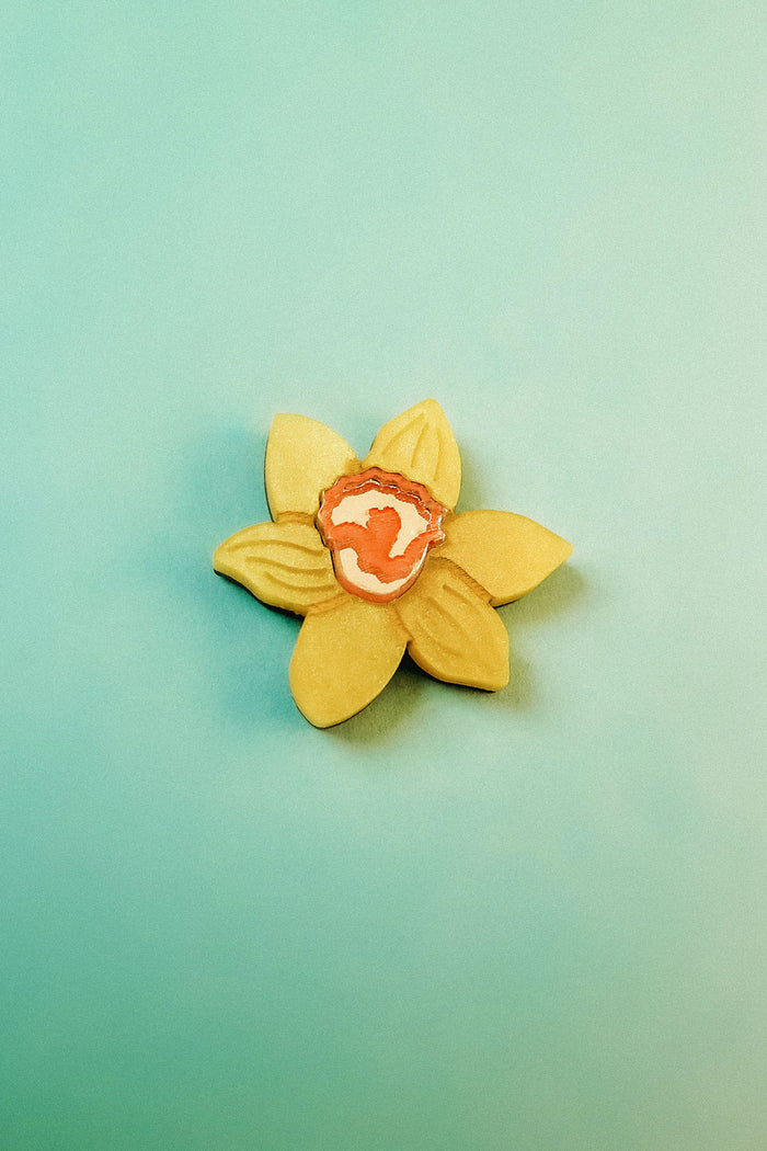 A handmade yellow flower-shaped cookie with an orange and white center is placed on a light teal background, reminiscent of a Daffodil Flower Lapel Pin crafted in acrylic.